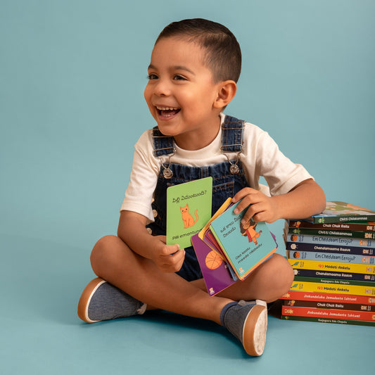 A young boy sitting cross-legged on a light blue studio background, holding colorful children's Telugu Flashcards. The flashcards have Telugu actions such as Oka Kaalu Meeda Nilabadu.