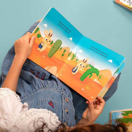 A child holding an open colorful children's book, Jinkundeluku Jemudante Ishtam with a desert scene featuring a Jinkundelu. Book is published by Bhaalu Books and is a Telugu board book written by Abhi Ilindra