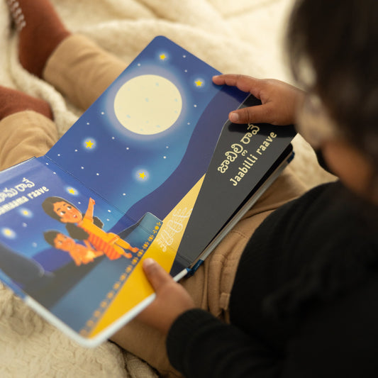 Child holding a telugu children's book chandamama raave with a night sky and moon illustration, sitting on a soft surface.
