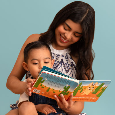 A woman and a young boy sitting on a light blue floor, reading a colorful children's book together. Book is Jinkundeluku Jemudante Ishtam by Abhi Ilindra. It is a Telugu-Engish bilingual board book.