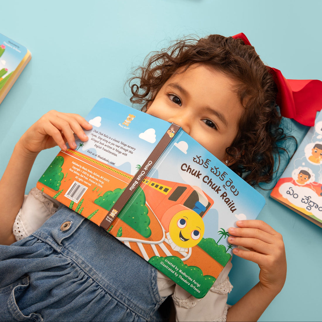 A young child with curly brown hair lies on a light blue surface, holding a colorful children's board book titled "Chuk Chuk Railu". It is a Telugu nursery rhyme made into a bilingual board book.