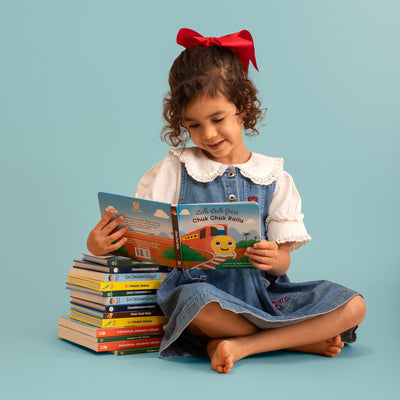 A young girl with curly brown hair and a red bow sits cross-legged reading Chuk Chuk Railu, a Telugu-English board book by Bhaalu Books. 